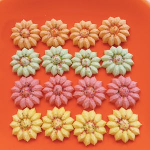 May include: A plate of 12 colorful sugar cookies decorated with sprinkles. The cookies are shaped like flowers and are arranged in three rows of four. The cookies are on a bright orange plate.