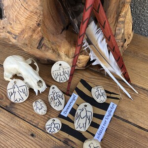 Peut inclure: Un ensemble de sept petits charmes en céramique blanche en forme d'oiseaux avec des dessins à l'encre noire. Les charmes sont disposés sur une surface en bois avec un papier rayé brun et noir avec le texte "Donnervogel schützt vor Unglück" à l'encre noire.