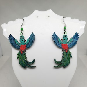 May include: A pair of wooden earrings featuring a vibrant Quetzal bird design. The birds have blue wings, red bellies, and long green tail feathers. The earrings hang from silver hooks with green beads.