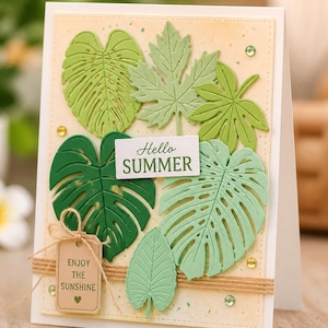 May include: A rectangular greeting card featuring die-cut paper tropical leaves in various shades of green. The card includes a white rectangular label with the text "Hello SUMMER" and a small tan tag tied with twine reading "ENJOY THE SUNSHINE".