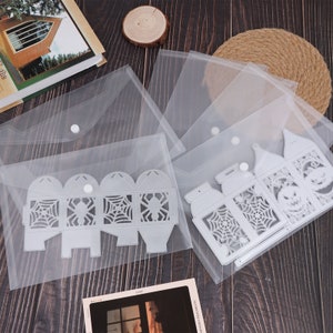 May include: Two clear plastic storage envelopes with metal die cut designs for crafting. The designs are of a lantern with a spider web and a jack-o-lantern.