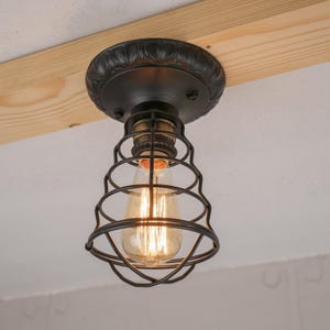 May include: A black, vintage-style ceiling light fixture with a cage-like shade. The fixture has a decorative, circular base and a visible Edison-style light bulb. The light is mounted on a wooden beam.