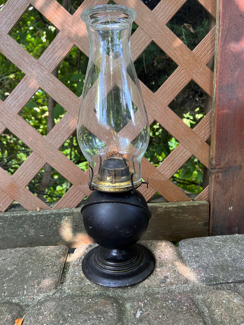 May include: A black oil lamp with a clear glass chimney. The lamp has a round base and a simple design.