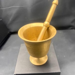 May include: A small, gold-colored mortar and pestle set. The mortar is a cup-shaped bowl with a flared rim and a round base. The pestle is a long, cylindrical rod with a rounded end.