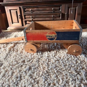 May include: A vintage wooden toy wagon with the Pepsi logo. The wagon features a red and blue painted design with the Pepsi logo in the center. The wagon has wooden wheels and a pull handle.