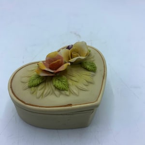 May include: A heart-shaped trinket box with a floral design. The box is made of ceramic and has a light beige color. The flowers are yellow, orange, and green.