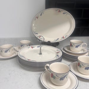 May include: A set of white ceramic dishes with a floral design in shades of blue, red, and green. The set includes a large oval serving platter, a cake stand, teacups, and saucers.