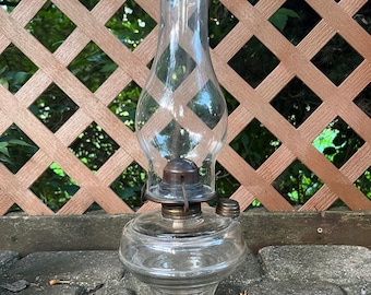May include: A clear glass oil lamp with a metal wick holder and a glass chimney. The lamp has a round base and a small handle on the side.