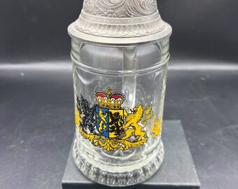 May include: A clear glass beer stein with a pewter lid featuring an ornate design. The stein has a colorful coat of arms with a crown and lions on it.