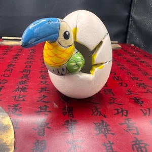 May include: A ceramic toucan figurine painted in bright colors, with blue, yellow, and green accents, emerging from a cracked white egg.