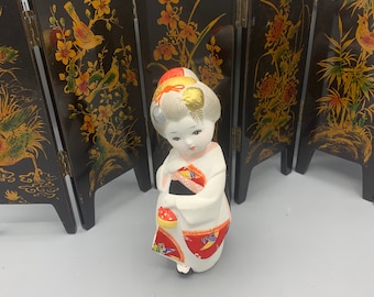 May include: A porcelain doll wearing a white and red kimono with gold accents. The doll is standing in front of a black folding screen with gold and red floral designs.