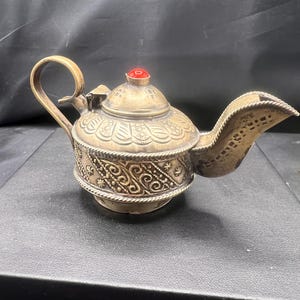 May include: A small, ornate silver teapot with a red jewel on the lid. The teapot has intricate floral and geometric designs.