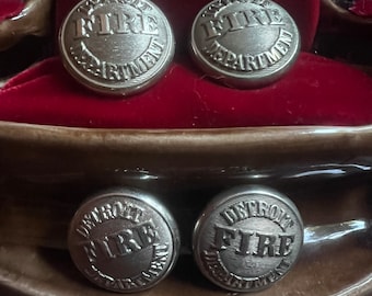 May include: Four silver metal buttons with the words "Detroit Fire Department" engraved on them. The buttons are arranged in two pairs, with the top pair on a red velvet surface and the bottom pair on a brown surface.