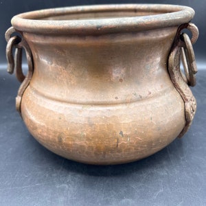 May include: A hammered copper planter with two ornate handles. The planter has a rounded bottom and a wide, open top.