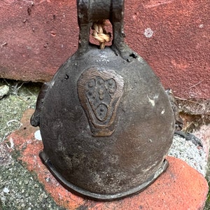 May include: An antique bronze bell with a decorative design on the front. The bell has a rope attached to the top.