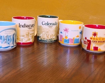 May include: Five ceramic mugs featuring different city designs. The mugs are white with colourful designs and text. The designs include Niagara Falls, Indianapolis, Colorado, Florida, and Las Vegas.