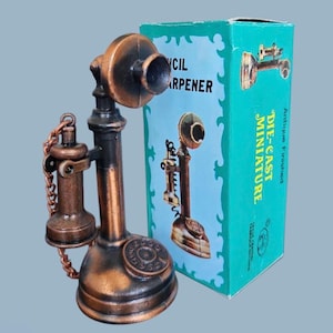 May include: A die-cast miniature antique telephone pencil sharpener. The pencil sharpener is bronze and black with a rotary dial and a receiver on a chain. The box is teal with the words "Pencil Sharpener" and "Die-Cast Miniature".