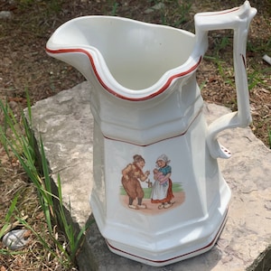 May include: White ceramic pitcher with a red rim and a handle. The pitcher features a painted scene of two people in traditional clothing.
