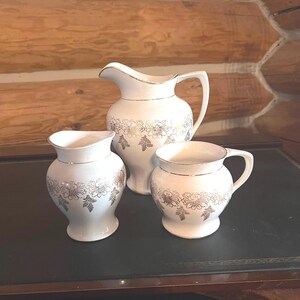 May include: A set of three white ceramic pieces with gold floral designs. The set includes a pitcher and two mugs.