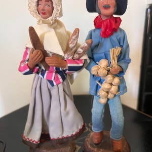 May include: Two handmade figurines, a woman in a white head scarf and a man in a blue shirt and black hat, holding loaves of bread and a bunch of garlic.