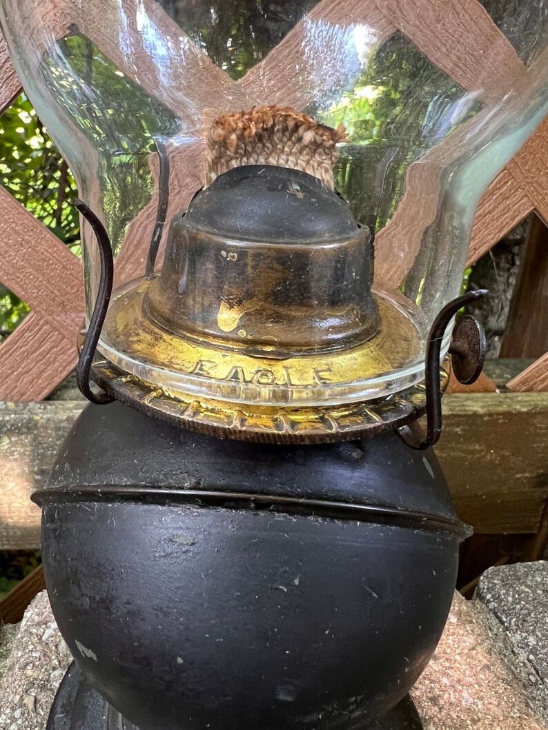 May include: Close-up of an antique oil lamp with a black metal base and a glass chimney. The lamp has a gold-colored metal top with the word "Eagle" inscribed on it.