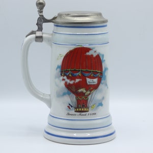 May include: A white ceramic beer stein with a pewter lid and a red, blue, and white hot air balloon design. The stein has a blue and white striped band around the bottom and the text "Benjamin-Marcel 7.5.1914" on the front.