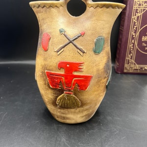May include: A brown ceramic vase with a handle and Native American designs. The vase features a red bird, crossed arrows, and footprints.