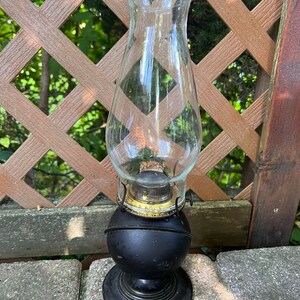 May include: A black oil lamp with a clear glass chimney. The lamp has a round base and a decorative gold band around the top.