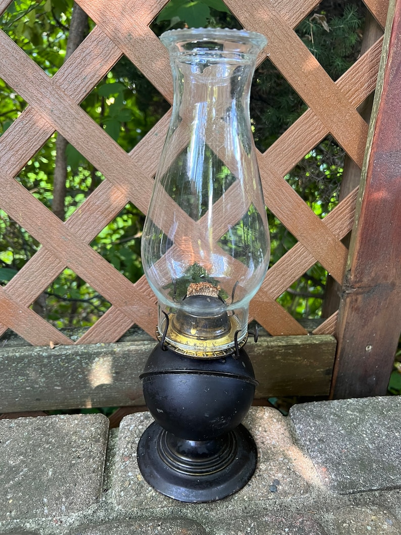 May include: A black oil lamp with a clear glass chimney. The lamp has a round base and a decorative gold band around the top.