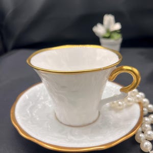 May include: A white teacup and saucer with a gold rim. The cup has a delicate floral design and the saucer has a matching design. A string of white pearls is lying next to the teacup and saucer.