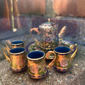 May include: A miniature tea set with a gold and blue teapot and four cups. The cups and teapot are decorated with floral patterns in pink, green, and gold.