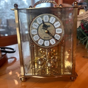 May include: A rectangular quartz clock with a gold-colored frame and a clear glass enclosure. The clock face is white with black Roman numerals and gold accents. A gold handle is at the top. The pendulum has multiple gold spheres.