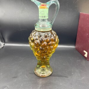 May include: A clear glass bottle shaped like a bunch of grapes, filled with a golden liquid. The bottle has a green handle and a label that reads "Pinord Chardonnay".