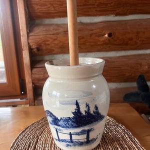May include: A cream-colored ceramic crock featuring a blue hand-painted landscape scene with a small house, trees, and a fence. A long, cylindrical wooden handle extends vertically from the center of the opening of the ceramic vessel.