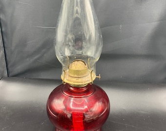 May include: A red glass oil lamp with a clear glass chimney. The lamp has a brass burner and a decorative beaded rim on the chimney.