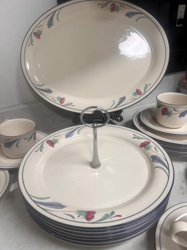 May include: A set of white ceramic plates with a blue and green floral design. The plates are stacked and include a cake stand with a silver handle.