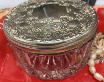 May include: A silver-plated round glass jar with a decorative lid. The lid has an intricate floral design. The jar is sitting on a red surface.