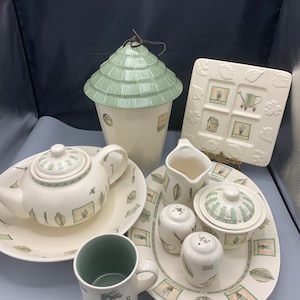 May include: A collection of cream-colored ceramic tableware with a garden theme. Includes a teapot, mug, serving tray, salt and pepper shakers, sugar bowl, creamer, and a decorative house-shaped container. The pieces are decorated with green accents and garden-related illustrations.