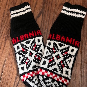May include: Pair of black wool socks with white and red geometric patterns. The socks have the word "ALBANIA" written in red on the top of each sock.