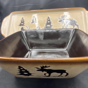 May include: Two-piece ceramic serving dish set. The set includes a square bowl and a rectangular dish. Both pieces are beige with brown rims and feature black silhouettes of trees and moose. The interior of the bowl is dark brown.