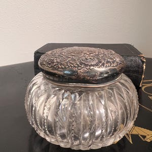 May include: A clear glass jar with a silver lid. The jar has a ribbed design and the lid has an ornate floral pattern.