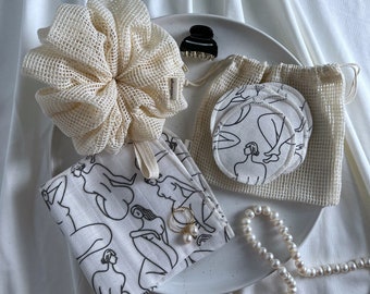 May include: A white ceramic plate with a white mesh scrunchie, a white mesh bag with round white cotton makeup remover pads, a white cotton cloth with a black line drawing of figures, a gold ring with a pearl, and a white pearl necklace.