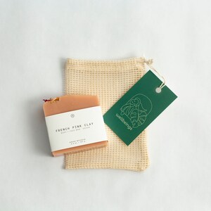 May include: A pink clay soap bar with a white label that reads "French Pink Clay Body & Face Bar with Kaolin". The soap bar is sitting on a beige mesh bag with a green tag that reads "wellbeings".