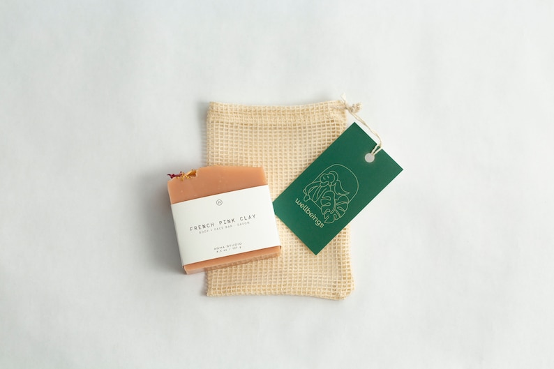May include: A pink clay soap bar with a white label that reads "French Pink Clay Body & Face Bar with Kaolin". The soap bar is sitting on a beige mesh bag with a green tag that reads "wellbeings".