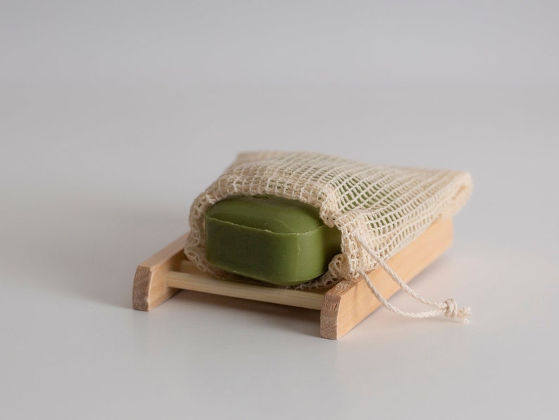 May include: A green bar of soap in a white mesh bag, sitting on a wooden soap dish.