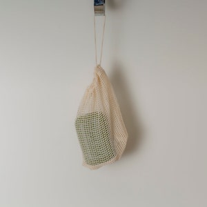 May include: A beige mesh produce bag hangs from a hook on a white wall. The bag is empty.