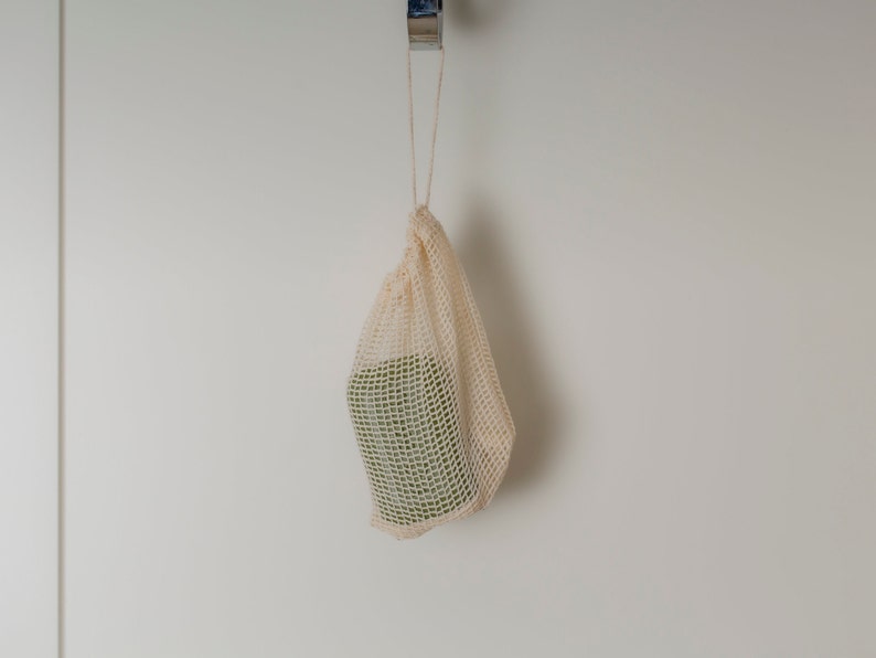May include: A beige mesh produce bag hangs from a hook on a white wall. The bag is empty.