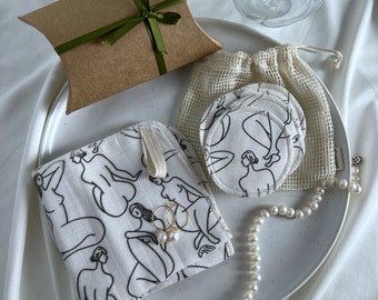 May include: A collection of eco-friendly beauty products and accessories. Includes a brown gift box with a green ribbon, a white plate with a patterned cloth, reusable cotton pads, and a pearl necklace. The cloth and pads feature a black line art design.