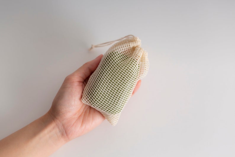 May include: A hand holding a small, off-white mesh bag with a green bar of soap inside.