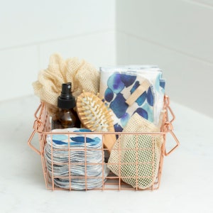 May include: A copper wire basket filled with bathroom essentials, including a reusable cotton round set, a natural hair brush, a mesh bag, a bottle of liquid, and a beige scrunchie.